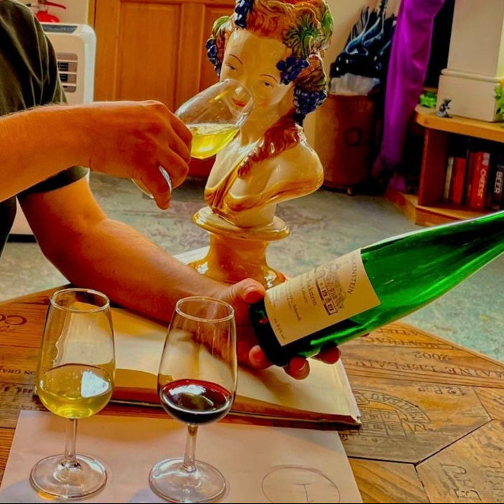 A person at a table holds a green wine bottle and pours white wine into a glass, with two wine glasses on the table and a decorative bust in the background.